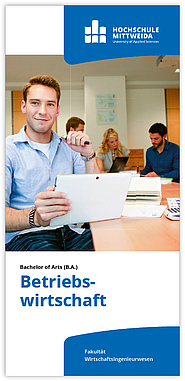 Ein junger Mann sitzt mit einem Tablet-PC am Konferenztisch. Im Hintergrund seine Kolleg:innen bei der Arbeit.